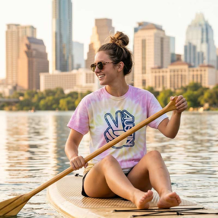 Woman on a paddleboard with a city skyline in the background wearing the Groovy Austin Desert Rose tie-dye t-shirt featuring a retro Austin peace sign graphic during a sunny day in Austin, Texas.
