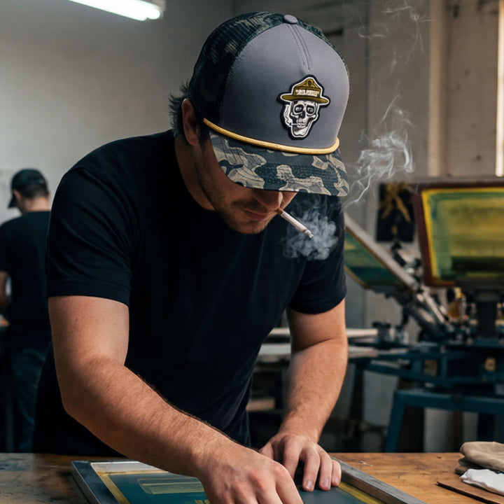 Person wearing the GTX Ranger Hat while working in a screen printing studio. Hat patch and topo camo brim visible in an authentic creative workspace.