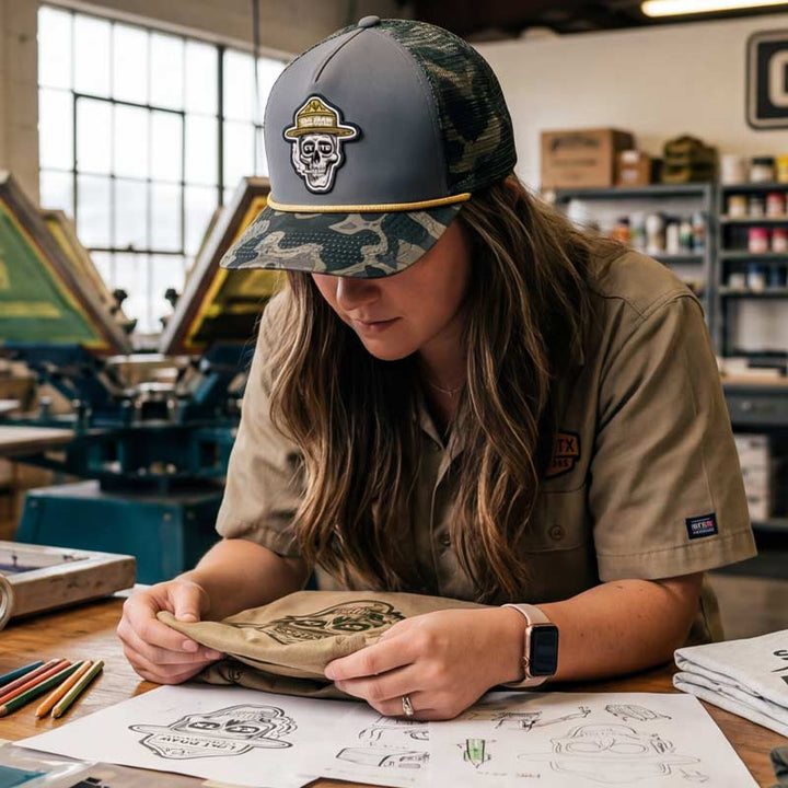 Woman wearing the GTX Ranger Hat while sketching designs in a print studio. Focus on the skull ranger patch and premium outdoor-inspired style in a creative Texas workspace.