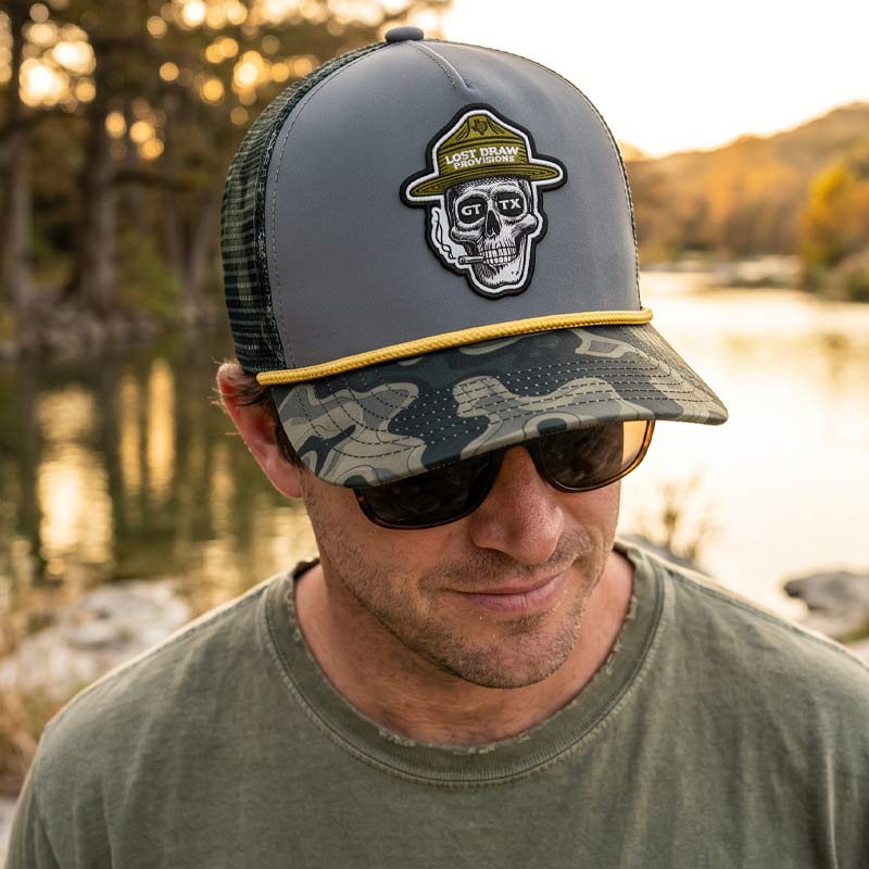 Man wearing the GTX Ranger Hat outdoors near a Texas river at golden hour. Skull ranger patch and camo bill visible with relaxed outdoor styling and warm natural light.
