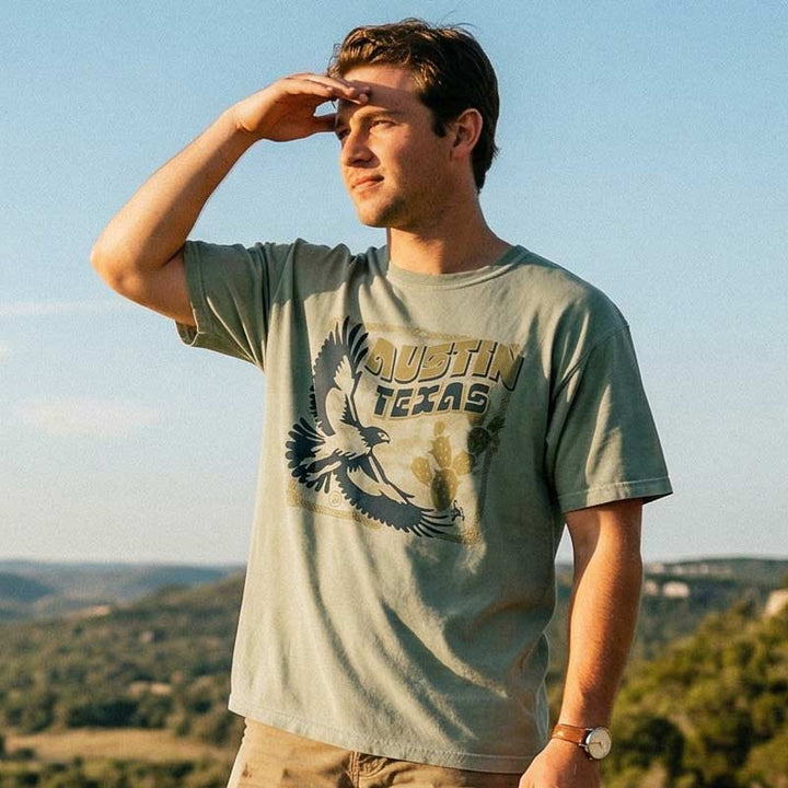 Man wearing an Austin Texas western graphic tee with hawk and prickly pear cactus design, standing outdoors with mountains in the background.