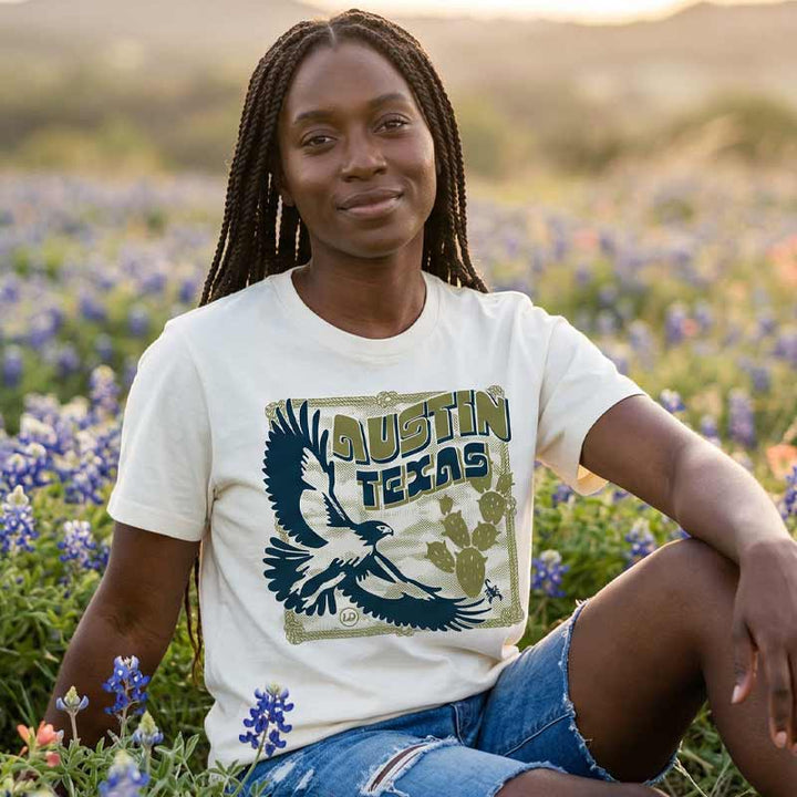 Women wearing heavyweight boxy tee with vintage hawk illustration in navy and olive tones in a field of blue bonnets.