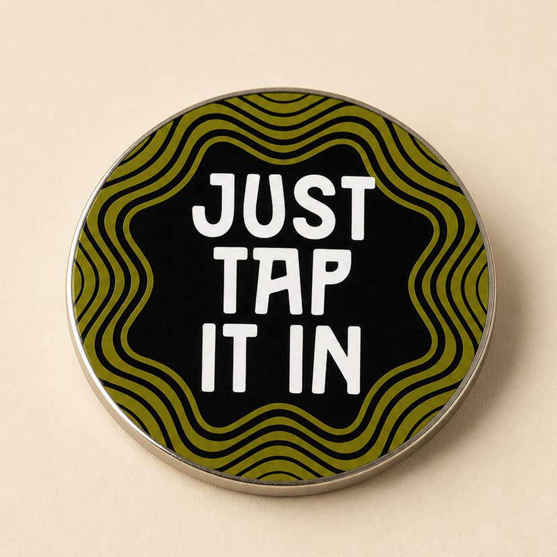 A round enamel pin with a black background, olive green wavy lines, and bold white text reading &