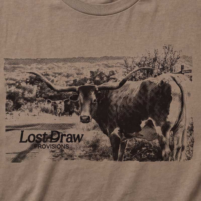 Close-up of a beige graphic t-shirt featuring a sepia-toned print of a longhorn cattle in a rural landscape with the text 'Lost Draw Provisions'.