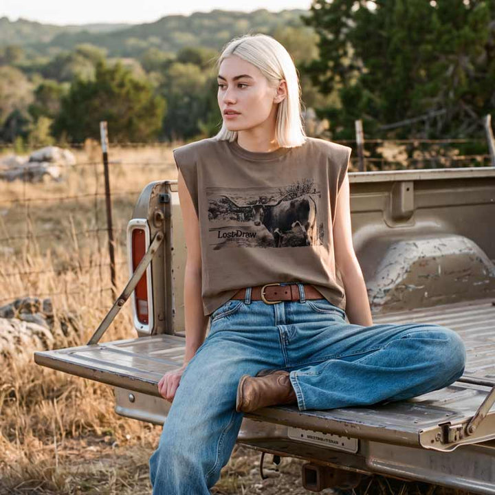 A woman with blonde hair sits in the bed of a pickup truck outdoors, wearing a sleeveless brown graphic t-shirt with a cow print and the text &