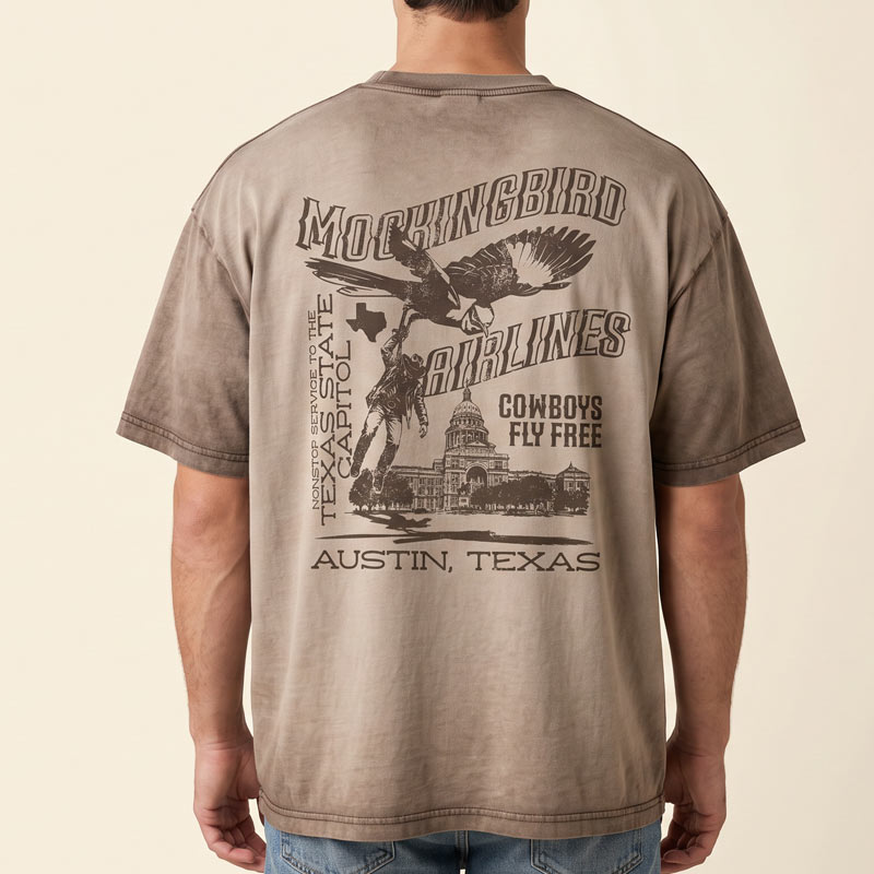 A man viewed from behind wearing a brownish-tan graphic t-shirt with a large black print of a mockingbird, cowboy, and Texas State Capitol on the back. The text reads &