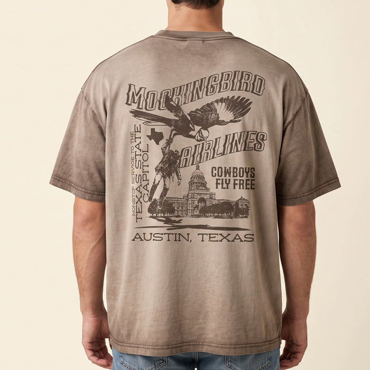A man viewed from behind wearing a brownish-tan graphic t-shirt with a large black print of a mockingbird, cowboy, and Texas State Capitol on the back. The text reads &