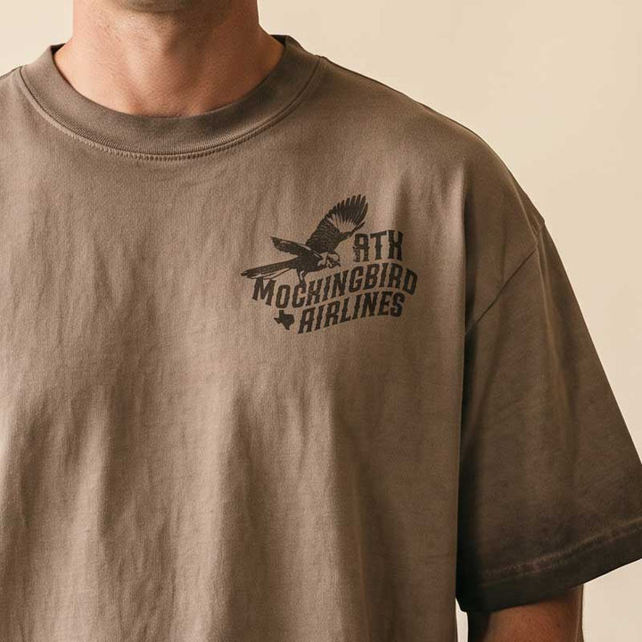 A close-up of a person wearing an oversized brown t-shirt with a black distressed graphic on the left chest featuring a mockingbird and the text &