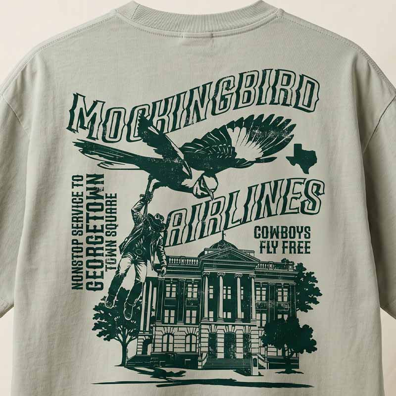 A close-up of a light beige graphic t-shirt showing the back print. The design features a dark green illustration of a cowboy riding a bird over a historic courthouse, with text reading &