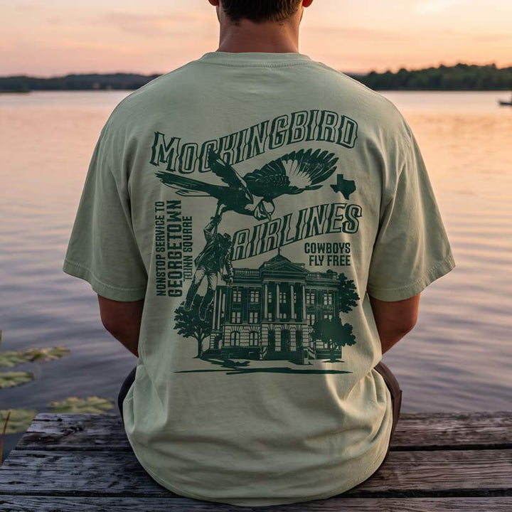 A man sitting on a wooden dock at sunset, viewed from behind, wearing a light sage green t-shirt with a large dark green graphic print on the back featuring the text &
