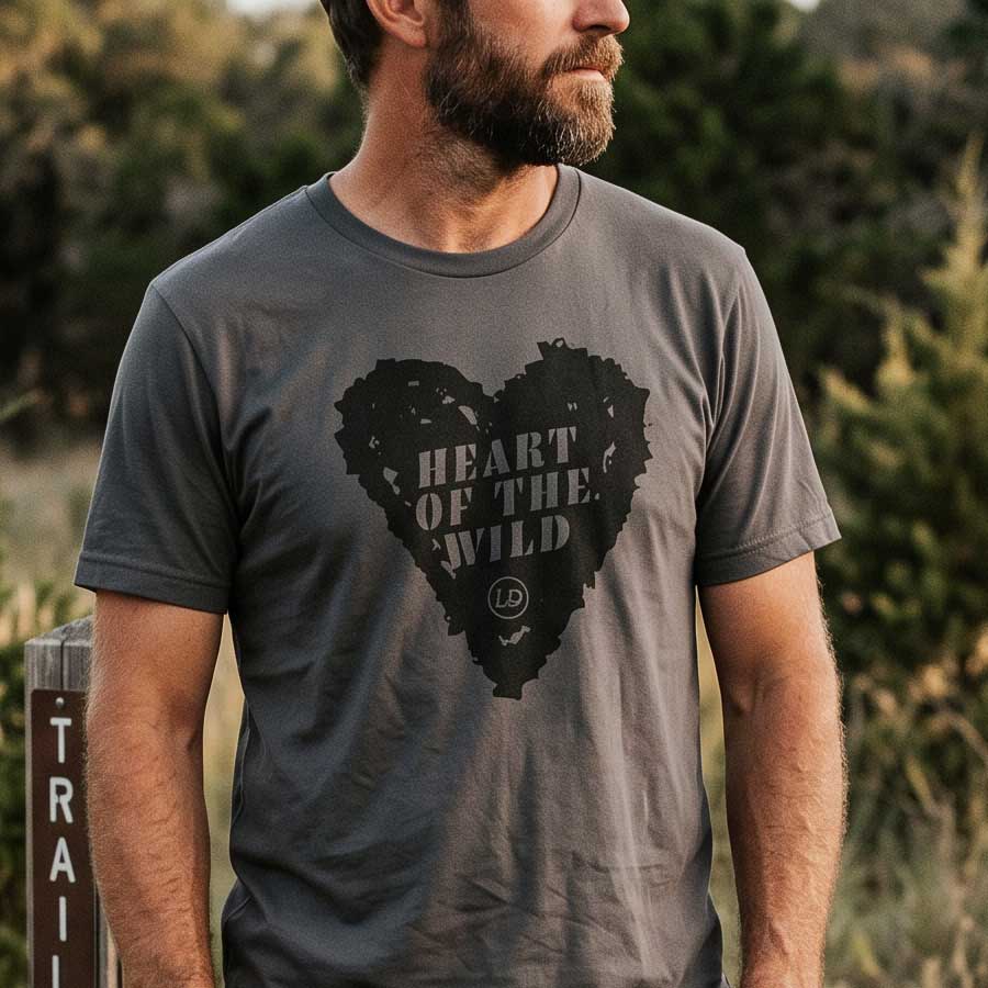A man wearing a dark gray crewneck t-shirt with a distressed black heart graphic that reads 'HEART OF THE WILD' and a small circular 'LP' logo, set against a blurred outdoor background.