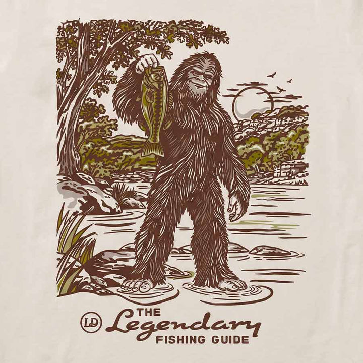 A close-up of a cream-colored fabric surface featuring a vintage-style graphic print of a Bigfoot-like figure holding a fish in a river, with the text &