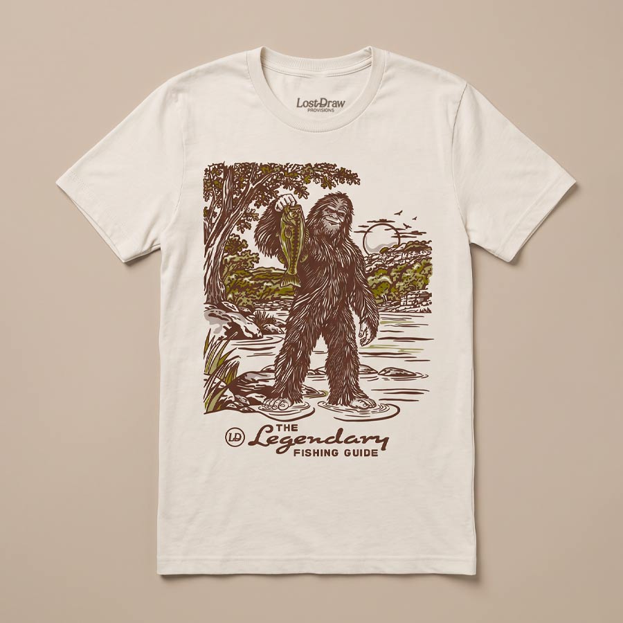 The Legendary Fishing Guide Tee in Vintage White featuring a vintage Bigfoot illustration holding a fish in a scenic river landscape.