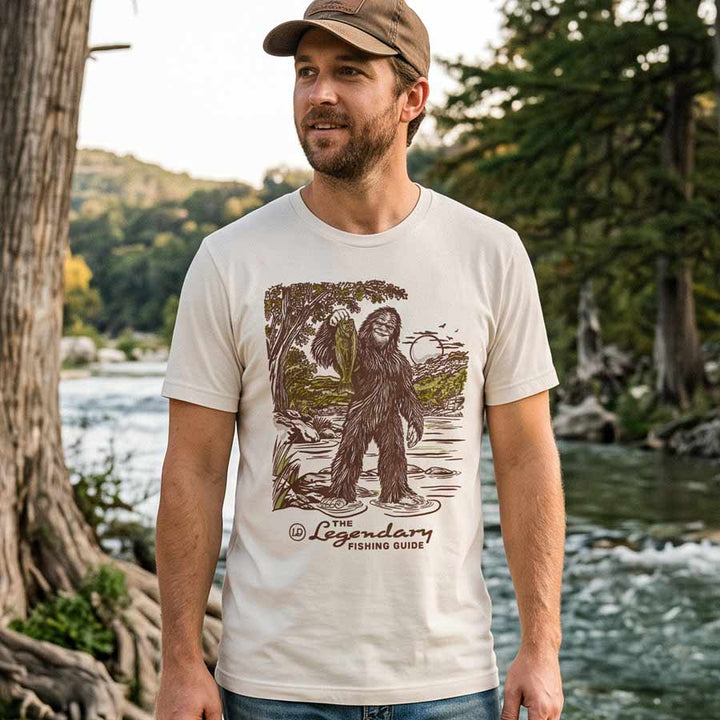 Male model wearing the Lost Draw Provisions Bigfoot fishing graphic tee in Vintage White beside a wooded river.