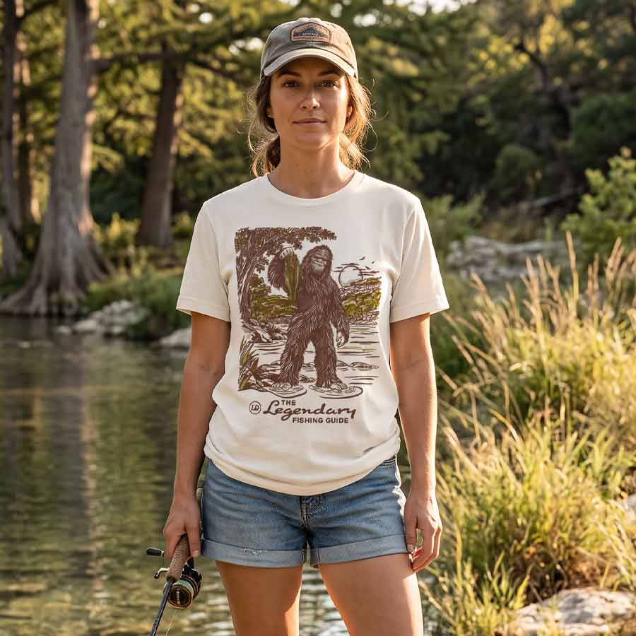 A woman stands outdoors by a lake wearing a cream graphic T-shirt with a large brown and green illustration of a creature and the text &