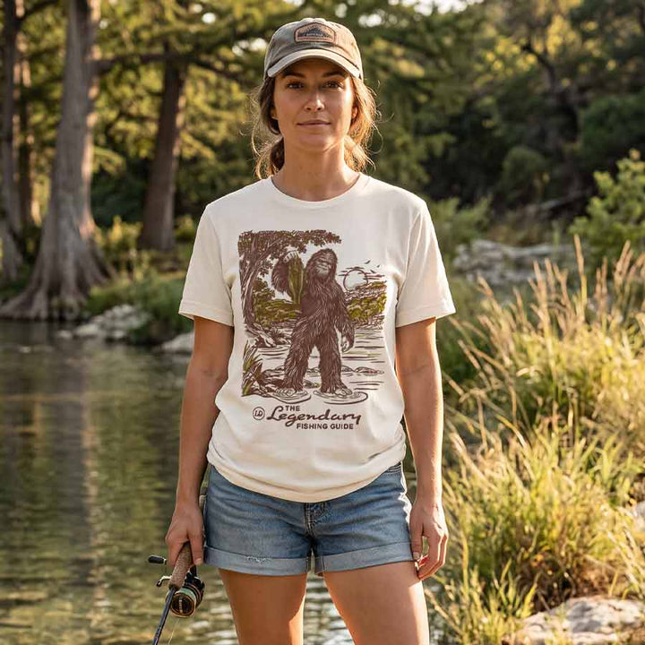 A woman stands outdoors by a lake wearing a cream graphic T-shirt with a large brown and green illustration of a creature and the text &