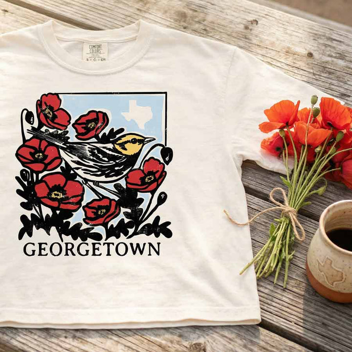 White t-shirt with bird and flower design on a wooden surface with flowers and a mug.