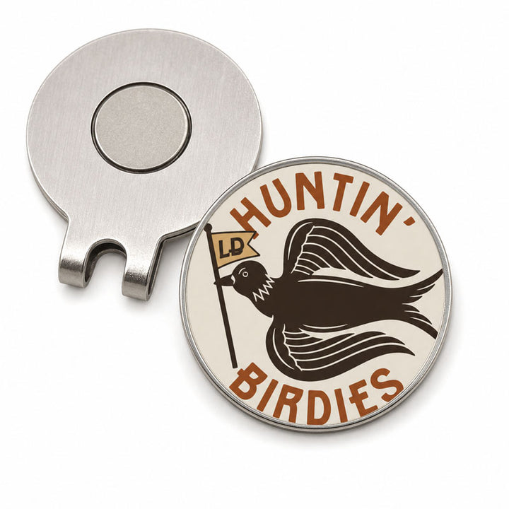 A silver metal magnetic golf divot clip shown next to a round engraved golf ball marker. The marker features a cream background with a dark brown bird holding a flag that reads &
