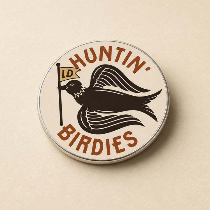 A round enamel pin with a silver metal border, featuring a black bird silhouette holding a small flag with &