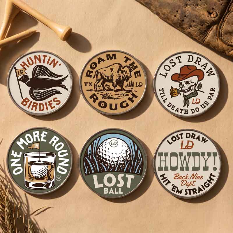 Six round golf-themed enamel pins arranged on a tan textured surface with rustic props including wooden tees and dried leaves. The pins feature vintage-style illustrations and text such as &