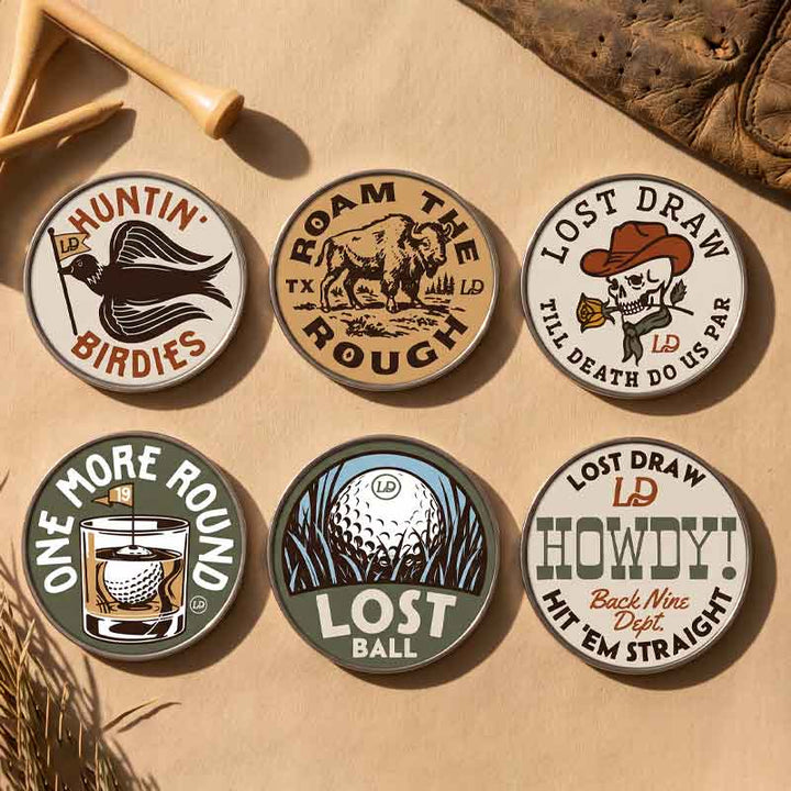 Six round golf-themed enamel pins arranged on a tan textured surface with rustic props including wooden tees and dried leaves. The pins feature vintage-style illustrations and text such as &