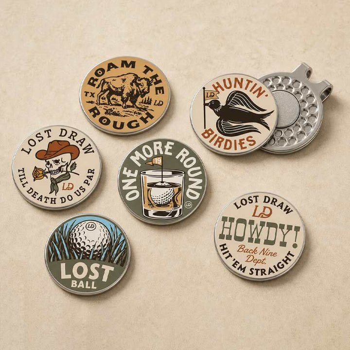 Six round golf-themed enamel pins arranged on a textured beige background, alongside a silver metal golf ball clip. The pins feature vintage-style illustrations and text such as &