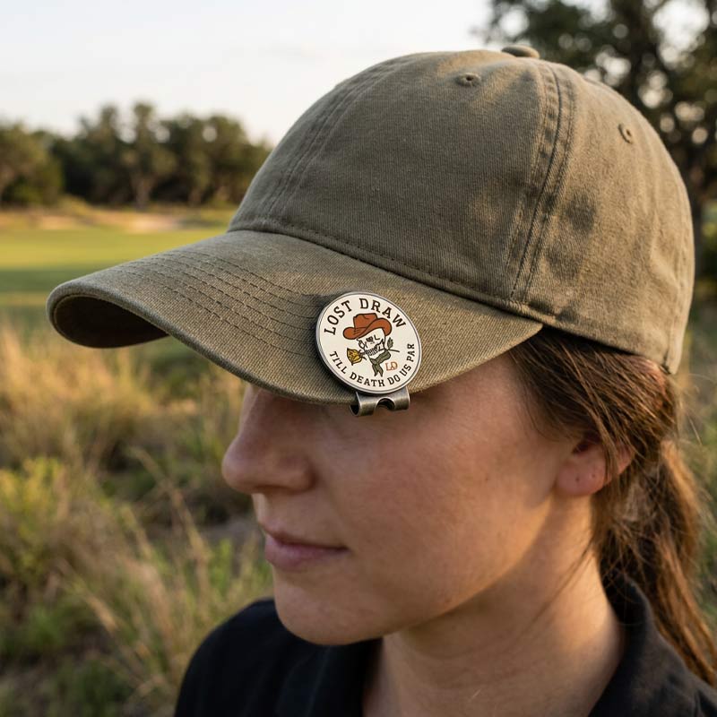A person outdoors wearing an olive green baseball cap with a round metal pin on the brim that reads &
