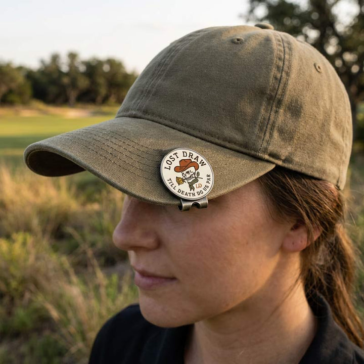 A person outdoors wearing an olive green baseball cap with a round metal pin on the brim that reads &