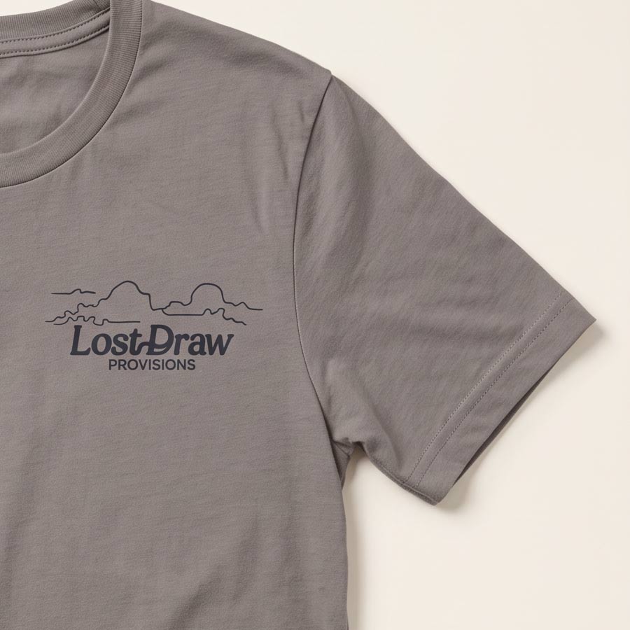 A close-up of a pebble brown short-sleeve t-shirt featuring a dark gray printed graphic on the left chest with the text &