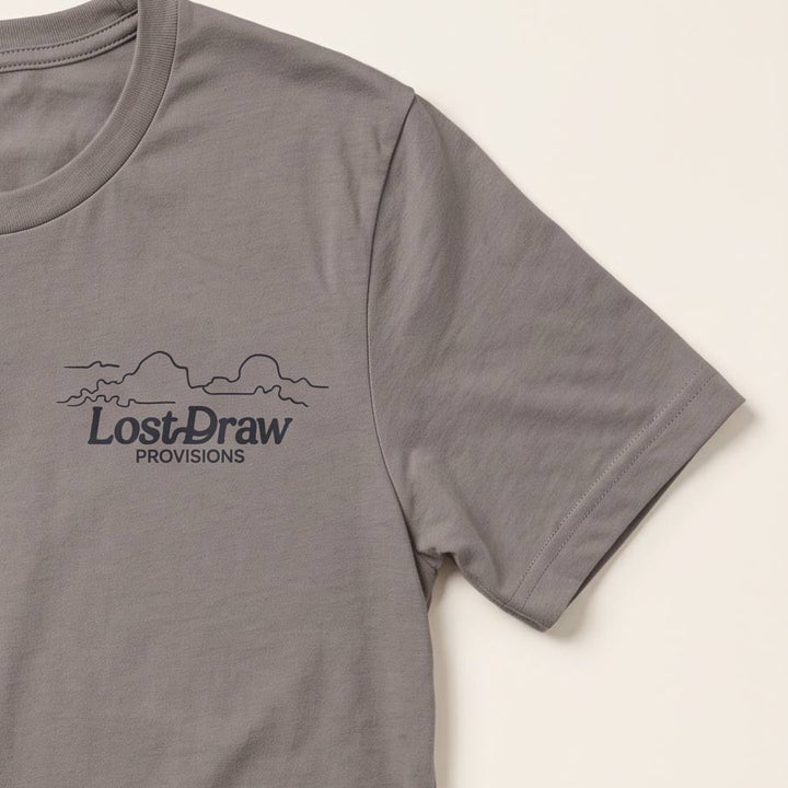A close-up of a pebble brown short-sleeve t-shirt featuring a dark gray printed graphic on the left chest with the text &