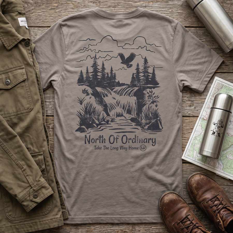Pebble Brown North of Ordinary Tee displayed with hiking boots, a canvas jacket, and a thermos, featuring the waterfall back graphic.