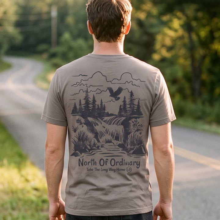 Man wearing the North of Ordinary Jersey Tee beside a tree-lined country road, displaying the waterfall and forest back graphic.