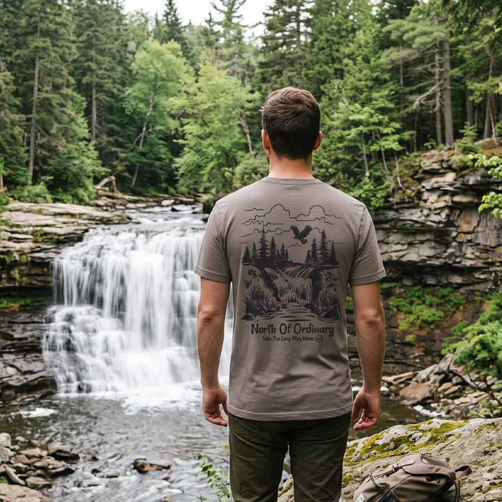 Man wearing the North of Ordinary Jersey Tee beside a waterfall, displaying the vintage outdoor graphic on the back.