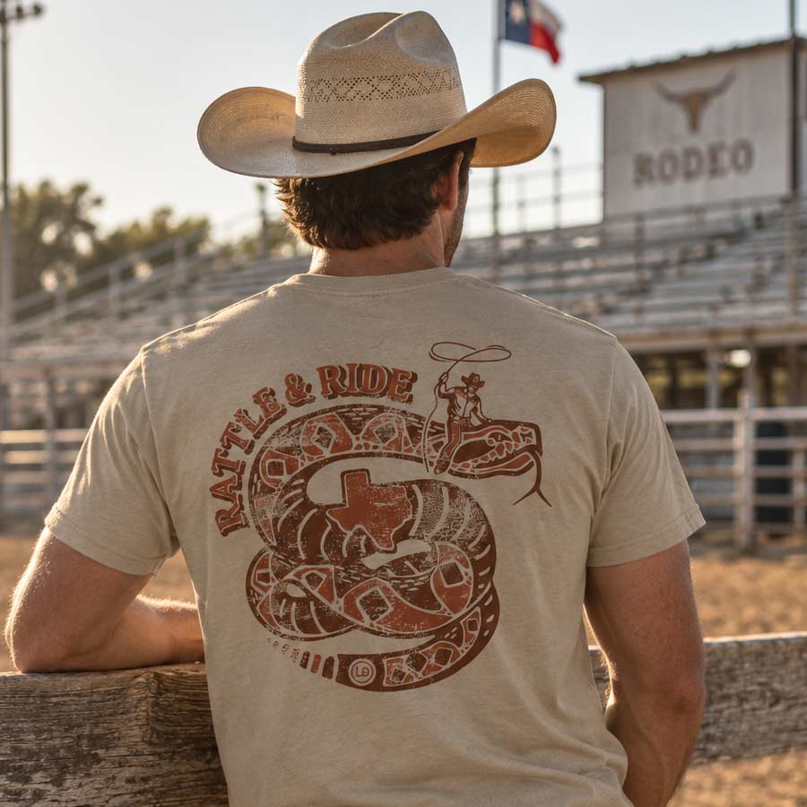 A man wearing a beige cowboy hat and a beige graphic t-shirt is seen from behind, sitting at a wooden fence in a rodeo arena. The back of the t-shirt features a large brown graphic of a coiled rattlesnake with a Texas state symbol and the words 'RATTLE & RIDE'.