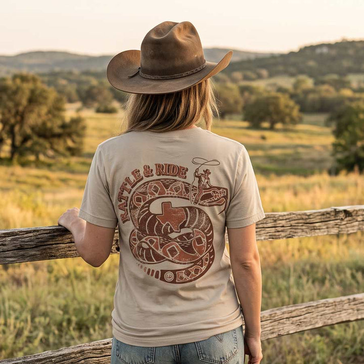 A woman viewed from behind wearing a tan cowboy hat and a beige graphic T-shirt with a large snake and &