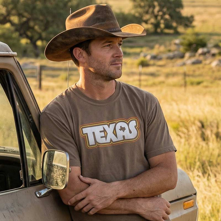 Man wearing a cowboy hat and 'Texas' t-shirt standing next to a vehicle in a field.