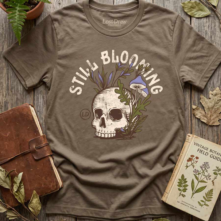 Vintage Brown Still Blooming graphic tee displayed with a potted fern, botanical book, and leather journal.