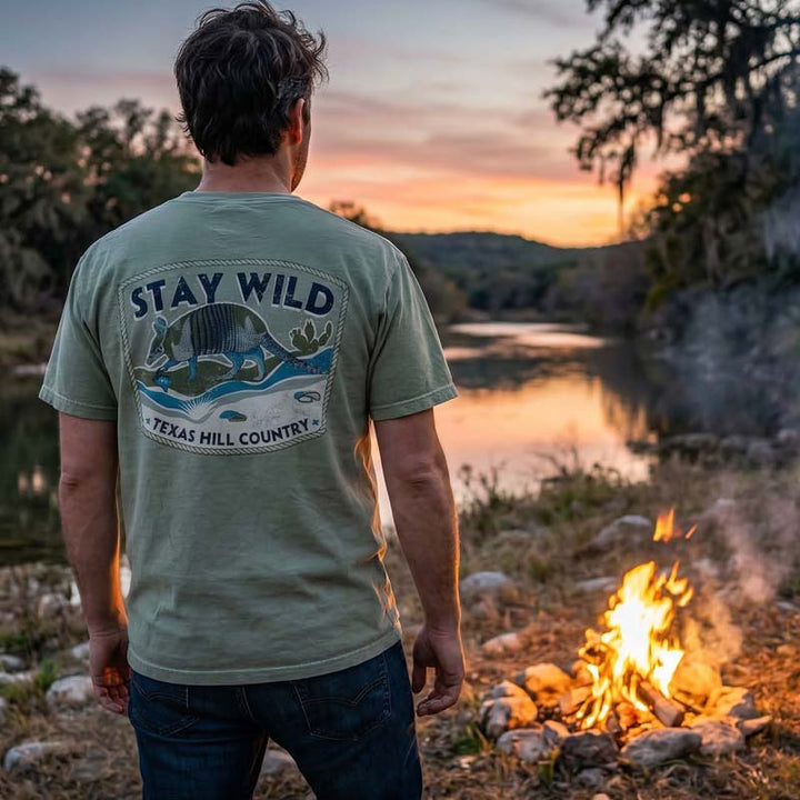 Person wearing a 'Stay Wild Texas Hill Country' t-shirt by a campfire with a scenic sunset.
