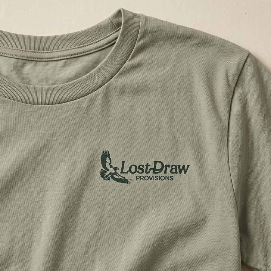 Details of Lost Hawk Jersey Tee in Thyme green featuring a small forest-green Lost Draw Provisions hawk logo on the left chest.