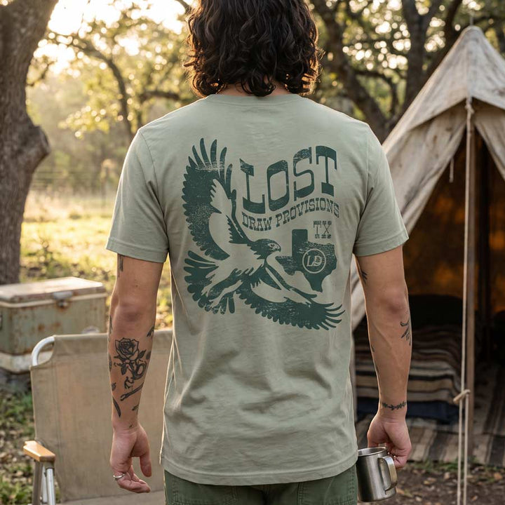 A man viewed from behind wearing an olive green t-shirt with a large dark green graphic on the back featuring an eagle, the word &
