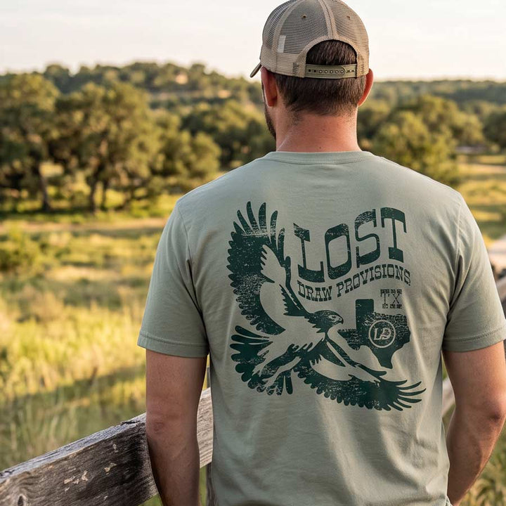 Back of the Lost Hawk Jersey Tee featuring distressed forest-green hawk illustrations, vintage Lost Draw Provisions lettering, and Texas artwork on a Thyme green shirt.