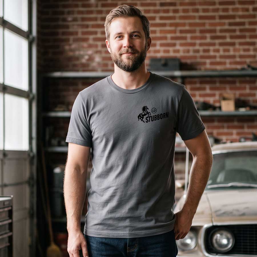 Man wearing the Asphalt Too Stubborn Jersey Tee inside a vintage garage, showing the small black horse and Stubborn chest graphic.