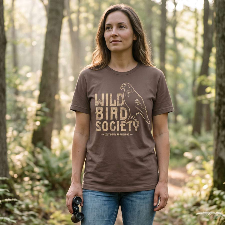 Woman wearing the Wild Bird Society Tee on a woodland trail, displaying the vintage bird illustration and cream lettering.