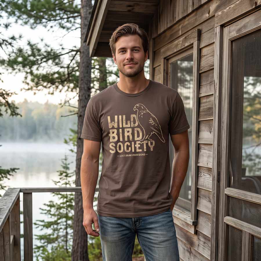 Man wearing the Cocoa Wild Bird Society Tee outside a lakeside cabin, showing the distressed bird graphic.