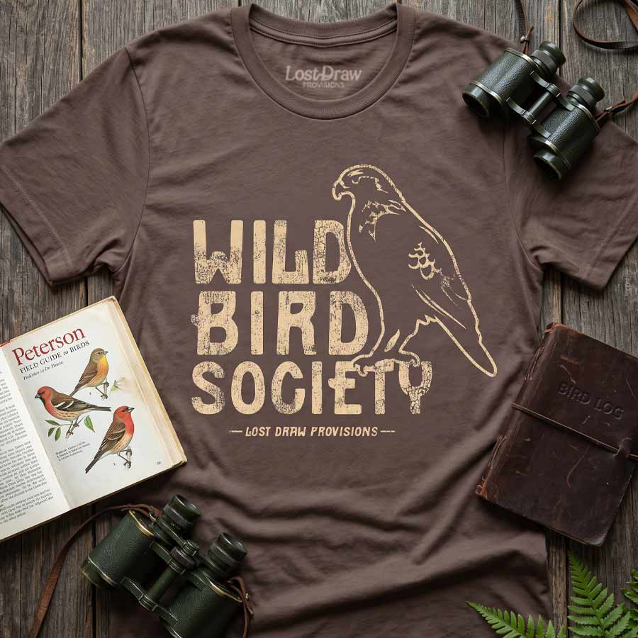 Cocoa Wild Bird Society Tee displayed with binoculars, a bird field guide, and a notebook, featuring the vintage cream bird graphic.