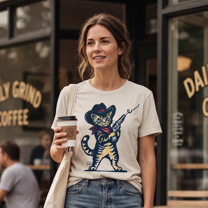 A woman wearing a beige graphic t-shirt with a cartoon cat in a cowboy hat holding a rifle, standing in front of a coffee shop window and holding a takeaway coffee cup.
